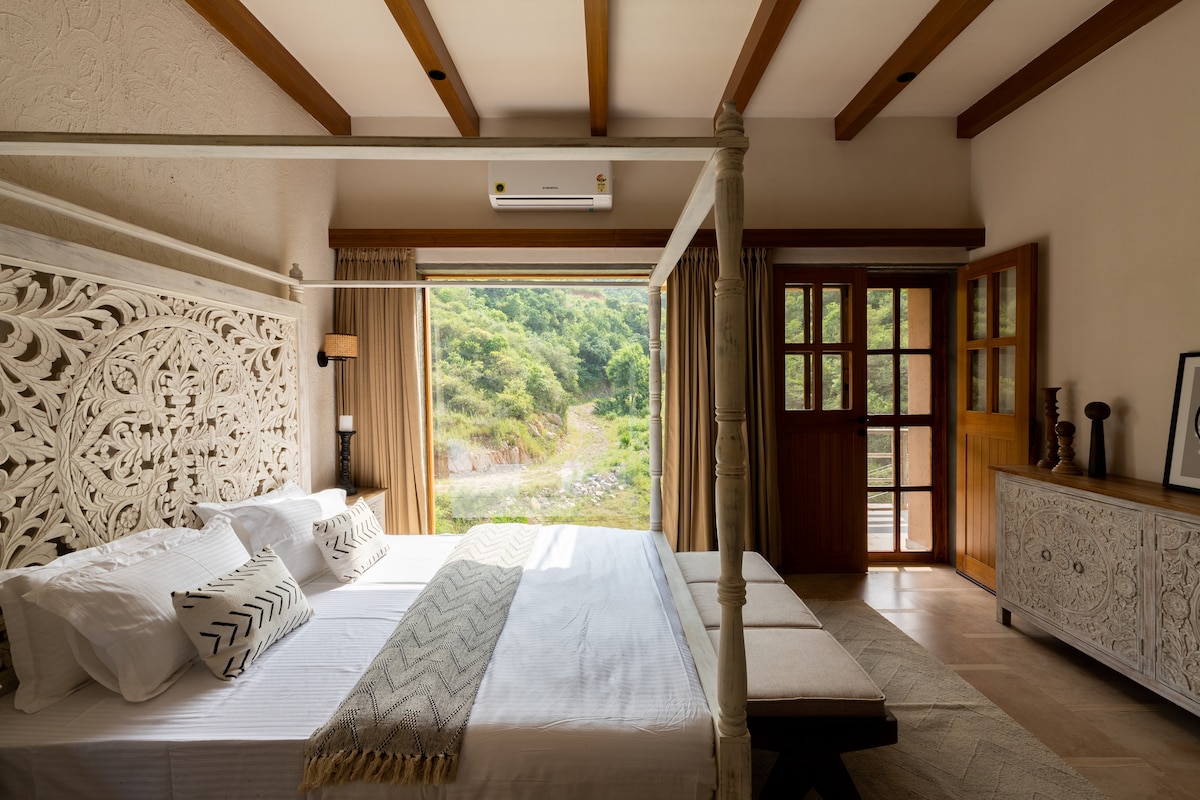 A bright bedroom features a four-poster bed adorned with intricate wooden detailing. Large glass doors open to a scenic view of rolling hills, allowing natural light to flood the space. A neutral palette and textured decor create a calming environment.