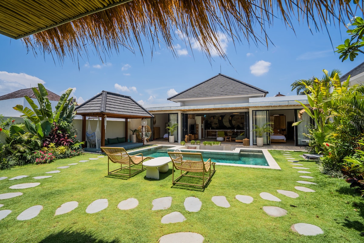An outdoor area features a refreshing pool surrounded by a well-kept lawn, with stepping stones leading to a gazebo. Two elegantly designed lounge chairs sit beside a circular table, while tropical plants provide a lush backdrop, creating a serene and inviting environment.