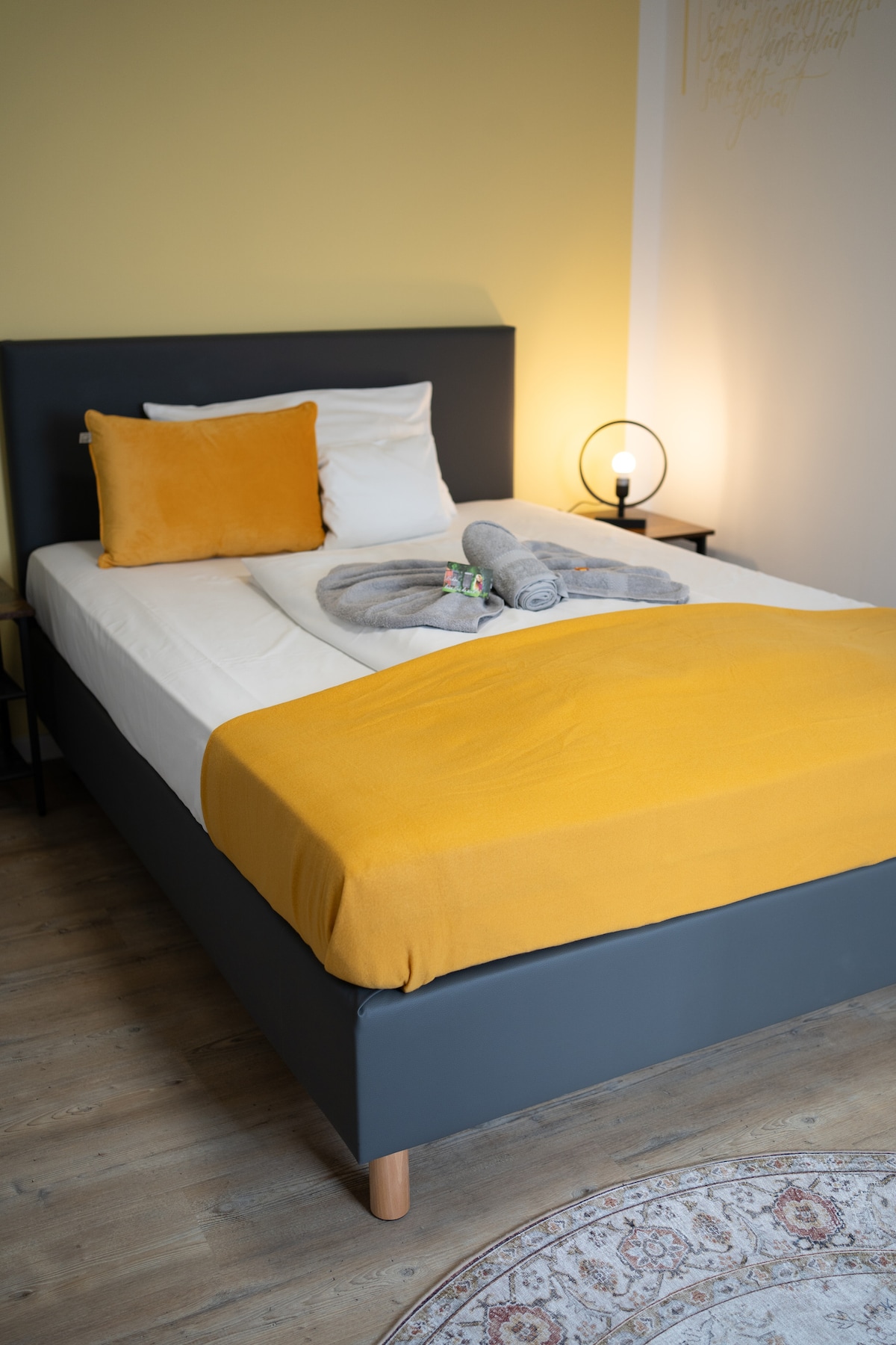 A minimalistic bedroom features a spacious bed covered with a yellow throw. Soft gray towels are neatly arranged on the bed, accompanied by a decorative accent pillow. A bedside lamp provides warm light, and the floor is adorned with a patterned rug.
