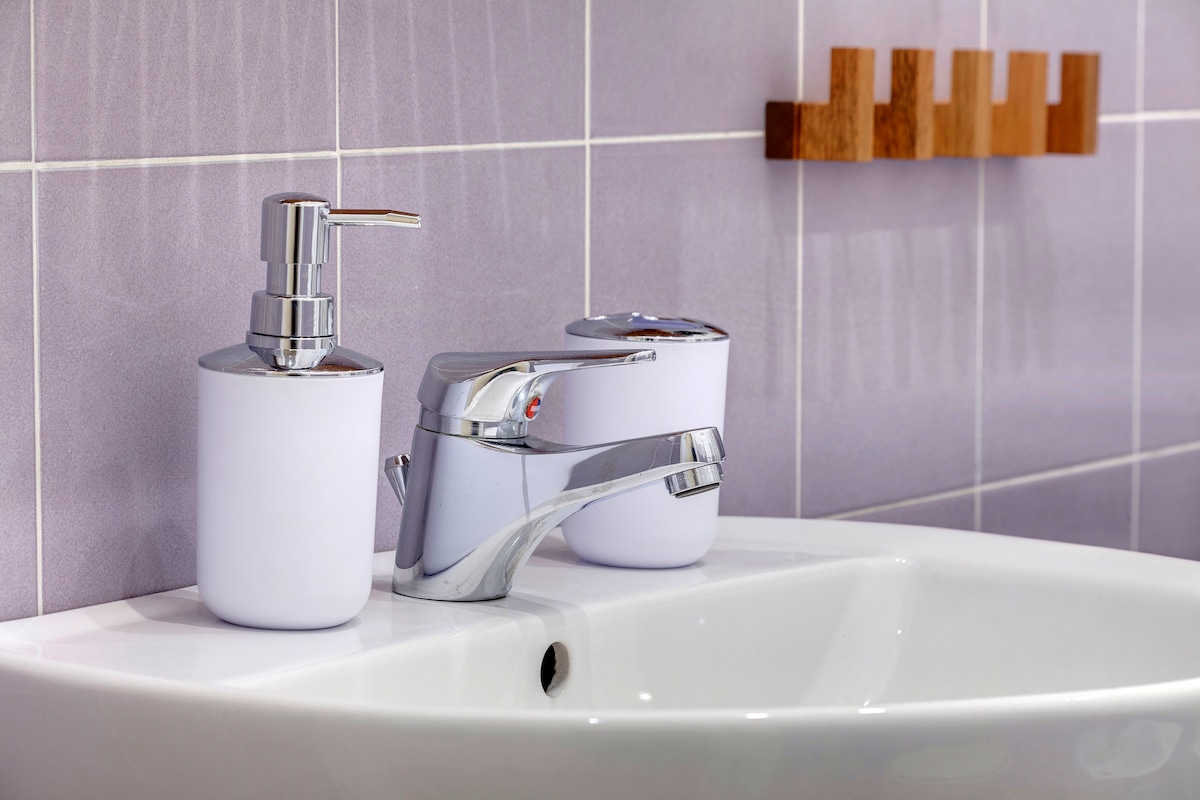 A modern bathroom sink is showcased with a sleek chrome faucet and a matching soap dispenser. The sink is surrounded by light purple tiles, while a wooden holder is mounted on the wall, providing space for toiletries.