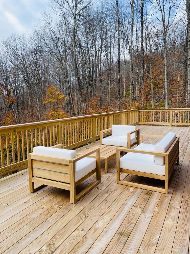 An expansive wooden deck is furnished with a cozy seating arrangement, featuring two armchairs and a coffee table. The backdrop showcases a serene wooded area, with bare trees and autumn foliage in warm hues, creating a peaceful outdoor retreat.