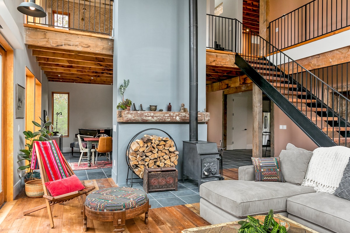 A spacious living area features a cozy gray sectional sofa and a decorative ottoman. A wood-burning stove and neatly stacked firewood add warmth, while large windows bring in natural light. A reclaimed wood staircase leads to an upper level, enhancing the open layout.