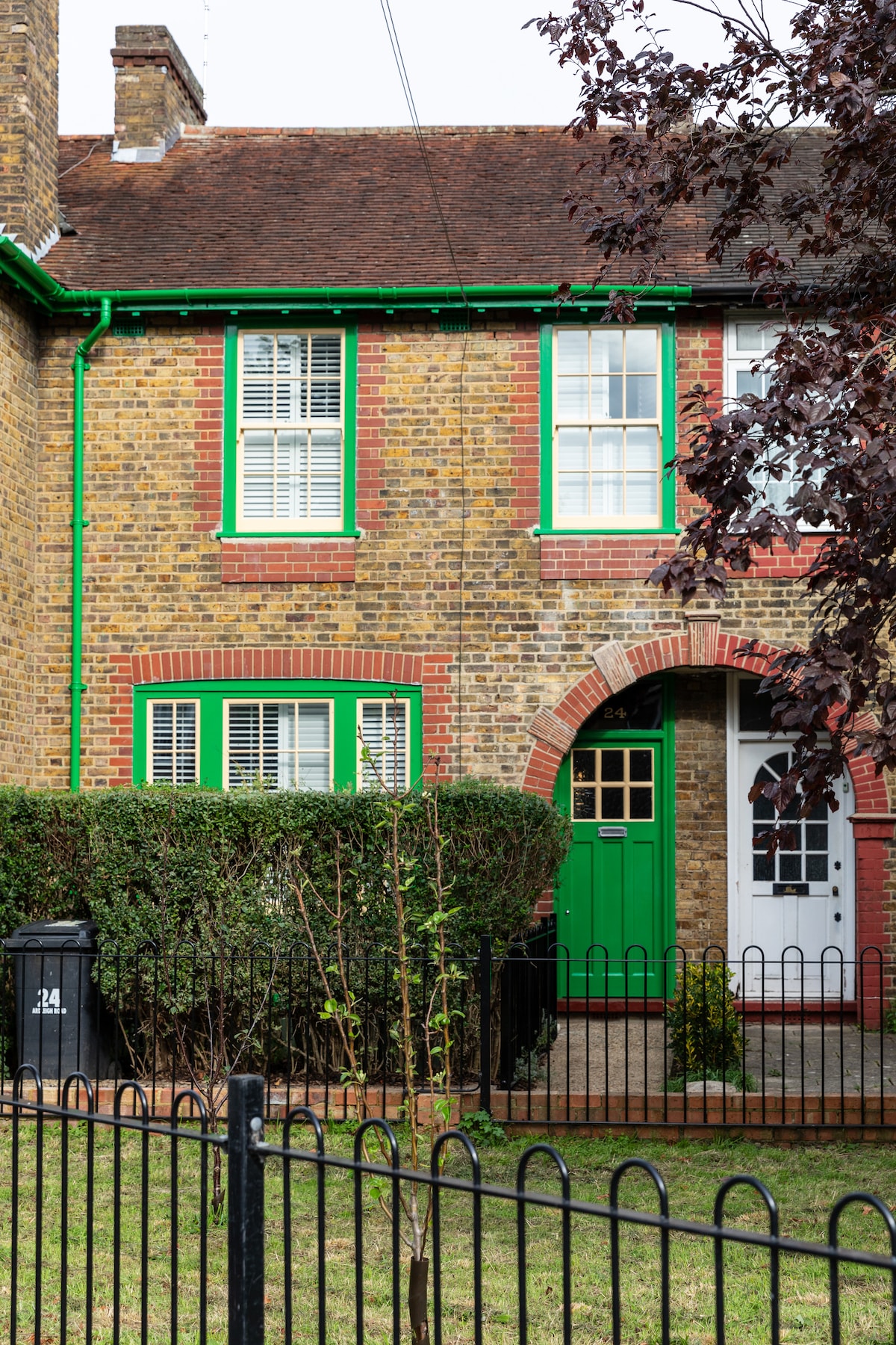A charming brick townhouse features bright green window frames and a matching front door. The entrance is adorned with decorative archways and surrounded by manicured hedges. A black recycling bin is positioned nearby, contributing to the overall tidy appearance of the front yard.