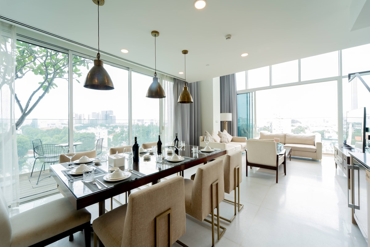 A bright and airy dining area is complemented by large windows offering views of the surroundings. A long table is set for dining, surrounded by soft beige chairs. Stylish pendant lights hang overhead, while comfortable seating is arranged nearby, creating a cohesive living space.