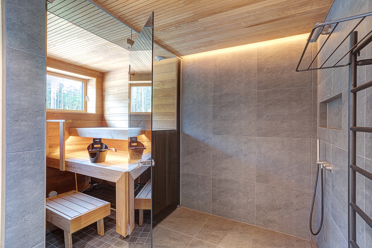 The sauna area features wood paneling complemented by sleek grey tiles. A modern rain shower with a glass partition is present, alongside a wooden bench. Natural light enters through a window, enhancing the serene and calming atmosphere.