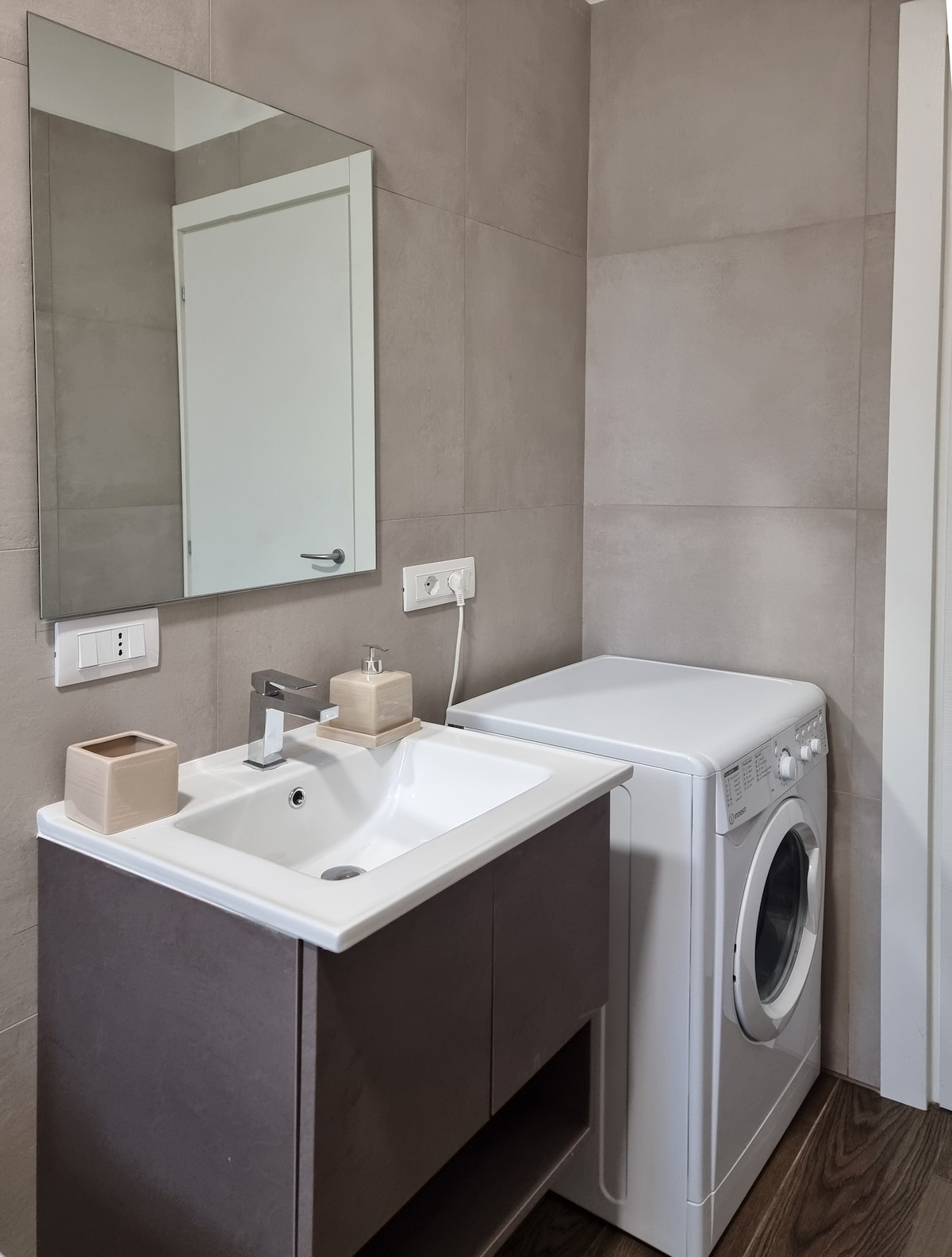 A compact bathroom features a modern sink with a sleek faucet and a large mirror above. A washing machine is positioned beside the sink, both placed against a neutral-colored wall. Light wood flooring adds warmth to the space.
