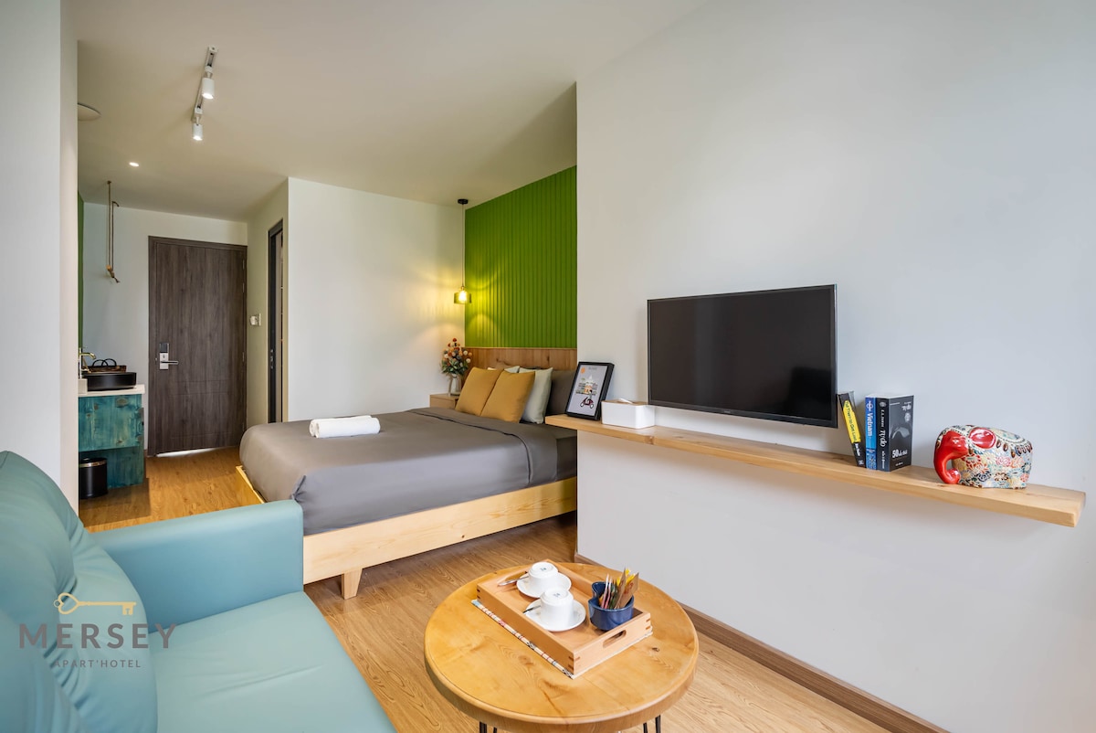 A stylish apartment room displays a spacious bed with neutral linens, backed by a vibrant green accent wall. A comfortable blue seating area is positioned beside a wooden shelf holding decorative items and a flat-screen television. Natural light filters through a nearby window.