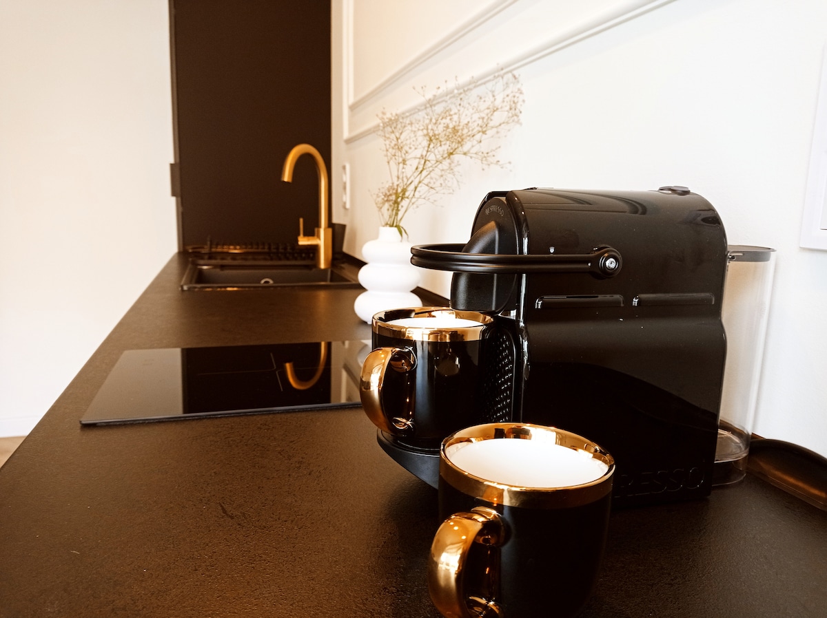 A modern kitchen area is showcased, featuring a sleek black countertop with a Nespresso machine and two elegant cups. A minimalistic white vase, containing dried flowers, adds a touch of sophistication against a contrasting dark wall.