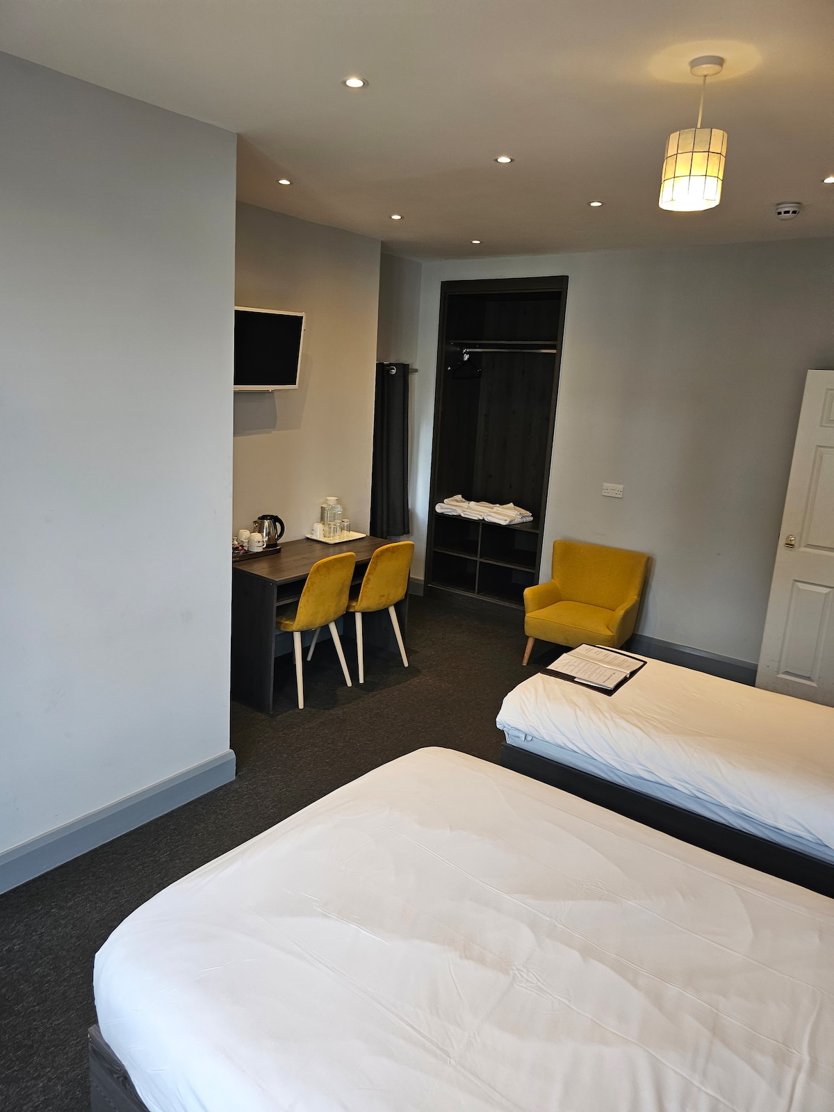A spacious room features two beds, a wooden desk with two yellow chairs, and a flat-screen TV mounted on the wall. Neutral-colored walls and ambient lighting create a clean environment. A wardrobe is visible, providing additional storage space.