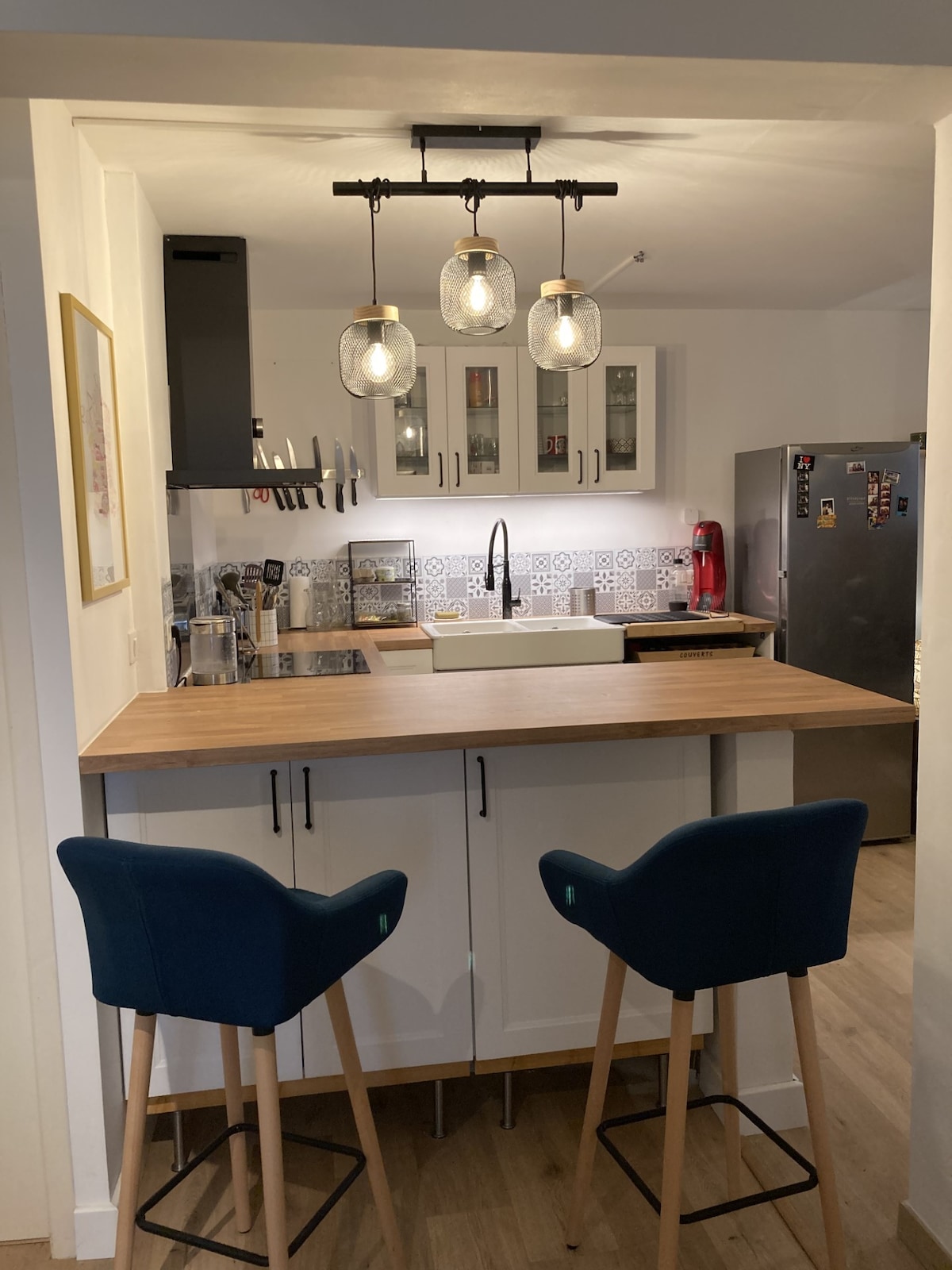 A modern kitchen area features a light wood counter with two blue bar stools. Above, three pendant lights provide illumination. The cabinetry showcases glass fronts, with a backsplash featuring intricate tile patterns. A stainless steel refrigerator stands in the background.