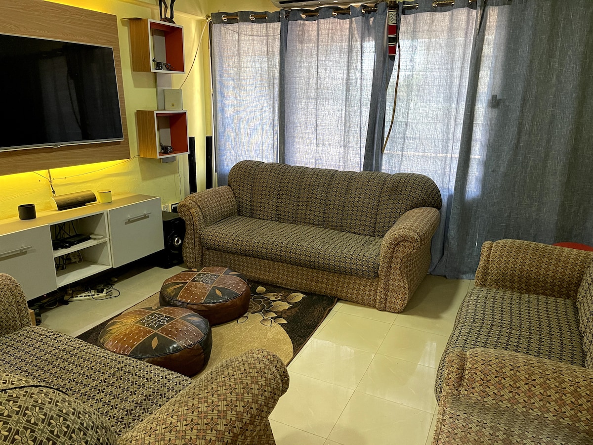 A cozy living area features two patterned sofas and a set of ottomans arranged on a patterned rug. Bright curtains frame a large window, allowing natural light to fill the space. A television is mounted on a stylish unit, providing an inviting setting for relaxation.