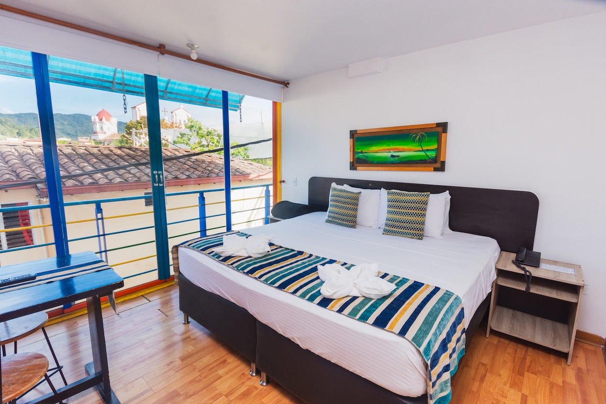 A spacious room features a double bed with crisp white linens and decorative pillows. Large windows provide natural light and offer views of the surroundings. A small table and two chairs are positioned beside the bed, enhancing the room's functionality.