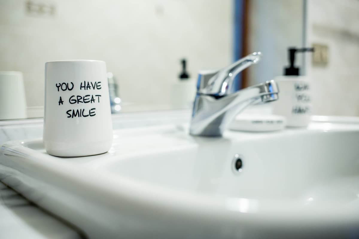 A clean bathroom sink is shown, featuring a white ceramic mug with the phrase 'YOU HAVE A GREAT SMILE.' A chrome faucet and a dark pump bottle are also visible. The scene reflects a simple and organized space.