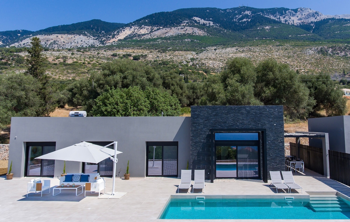 A modern villa is shown with a private pool surrounded by sun loungers and a shaded seating area. Large glass doors lead into the interior, which contrasts with the natural landscape of mountains and greenery in the background.