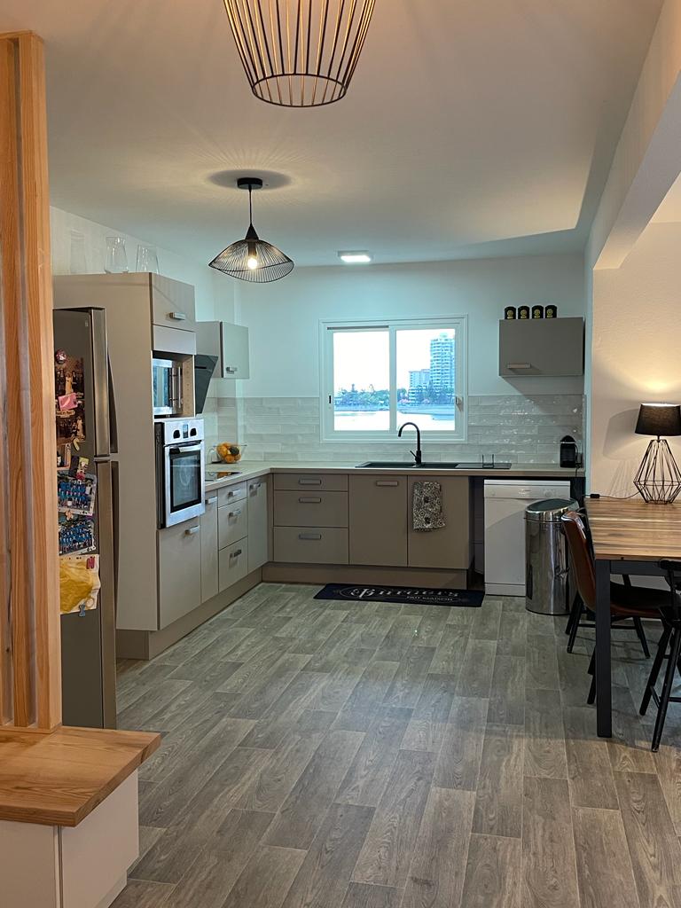 The kitchen features modern cabinetry with light tones and a well-equipped cooking space. Stainless steel appliances, including an oven and refrigerator, are present. A small dining table sits adjacent to a bright window, offering a view of the surroundings while enhancing natural light.