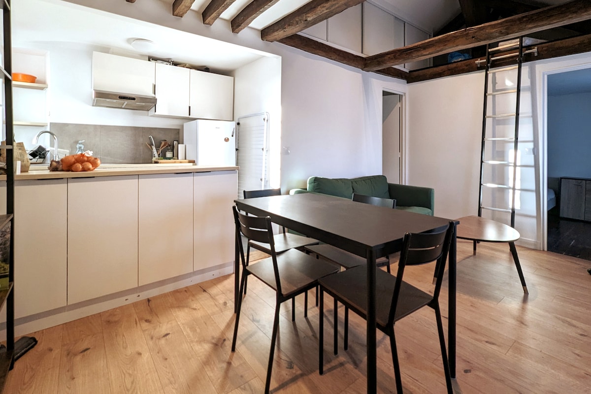 The open living area features a compact kitchen with modern appliances, a table with four chairs, and a cozy green sofa. Natural light fills the space through a window, and wooden beams add character to the ceiling. A ladder leads to an upper loft area.