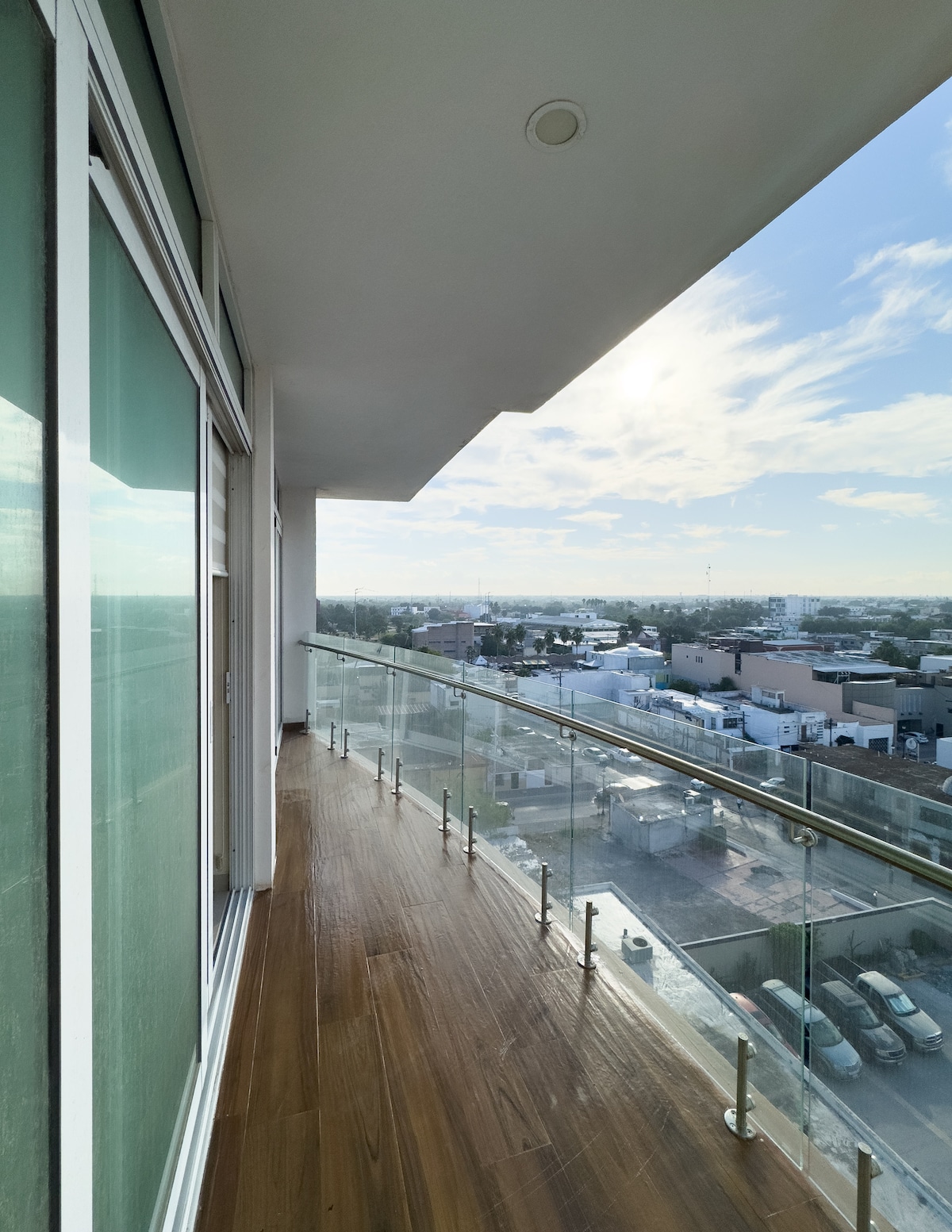 A spacious balcony with glass railing offers an unobstructed view of the surrounding area. The wooden flooring extends throughout, enhancing the open feel. Natural light fills the space, complemented by a clear blue sky and wispy clouds above.