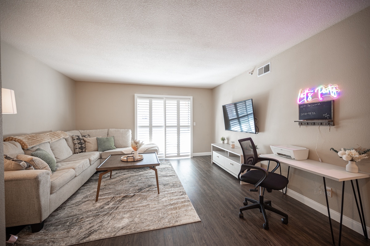 A spacious living area is furnished with a light-colored sectional sofa, a wooden coffee table, and a simple desk. A wall-mounted television is visible, along with a neon sign that adds a playful touch. Natural light filters through the window with white shutters.