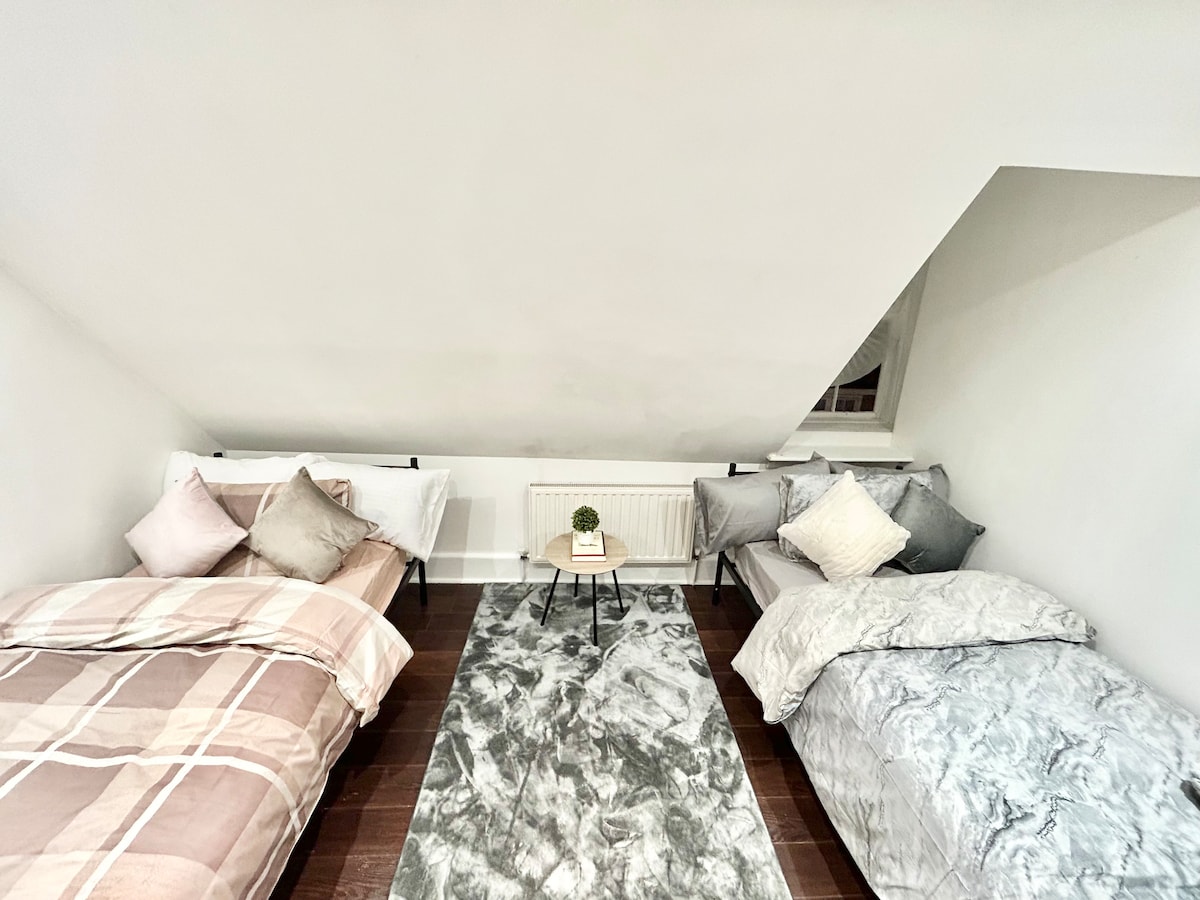 A bright sleeping area features two spacious beds covered with soft bedding in neutral tones. A small table sits between the beds, adding functionality, while a textured area rug introduces a cozy element to the hardwood floor.