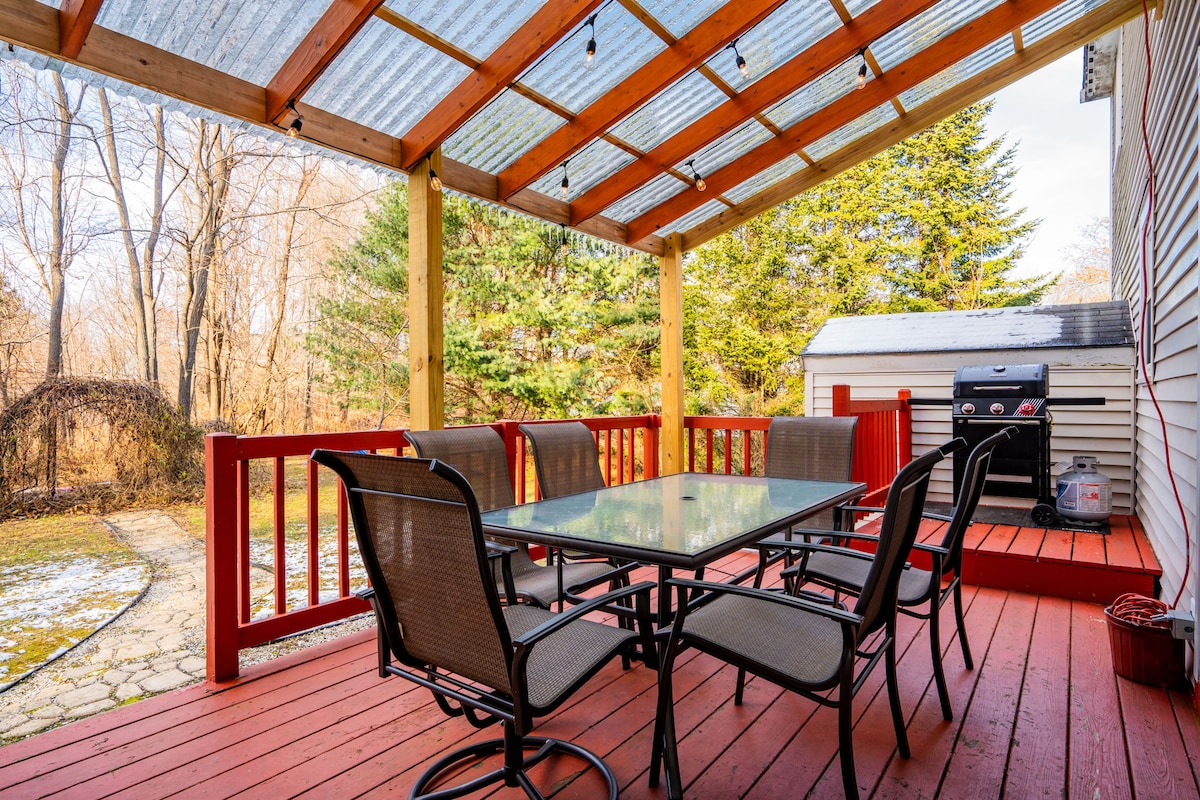 The covered deck features a spacious table with seating for six, surrounded by six chairs. A gas grill is positioned nearby. The area is illuminated by string lights above, while trees and greenery create a peaceful backdrop.