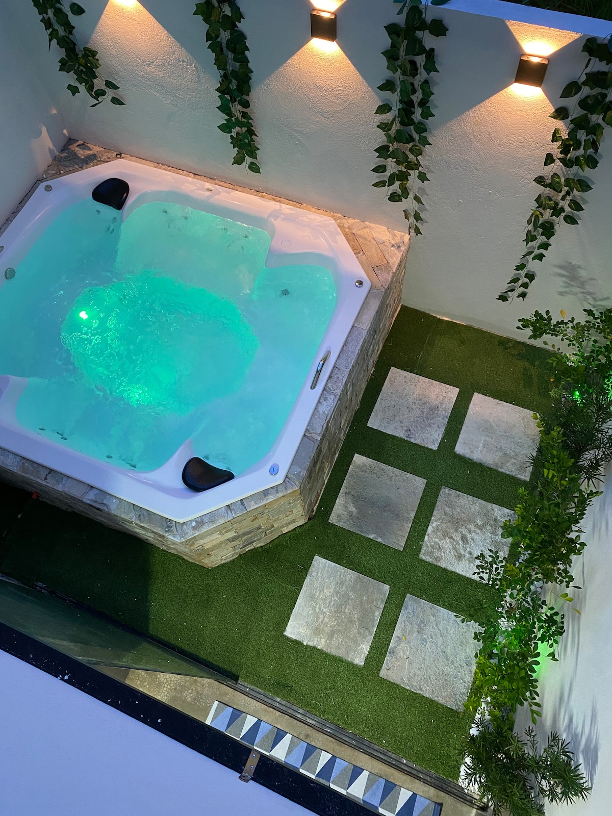 A private jacuzzi is set in a serene outdoor space, featuring gentle lighting that highlights its turquoise waters. Surrounding the jacuzzi are green artificial grass and neatly arranged stone pavers, creating a tranquil ambiance for relaxation.
