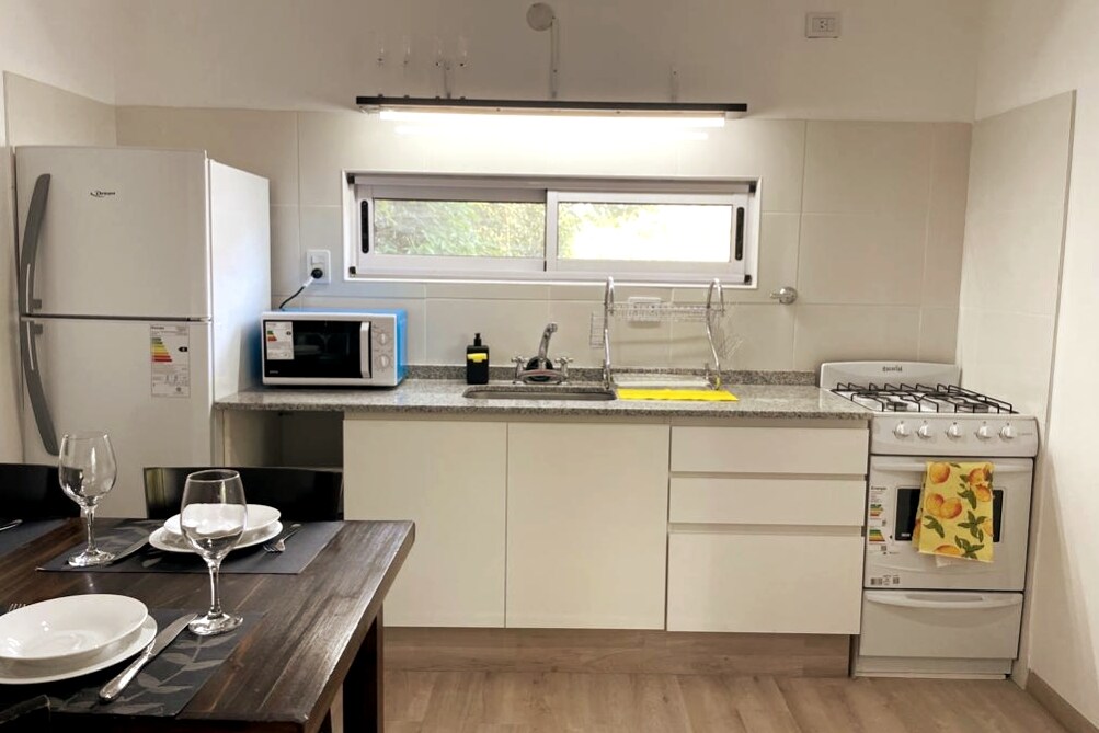 A functional kitchen space features white cabinetry and a granite countertop. Essential appliances include a refrigerator, microwave, and gas stove. The area is complemented by a dining table set with plates and glasses, and natural light enters through nearby windows.