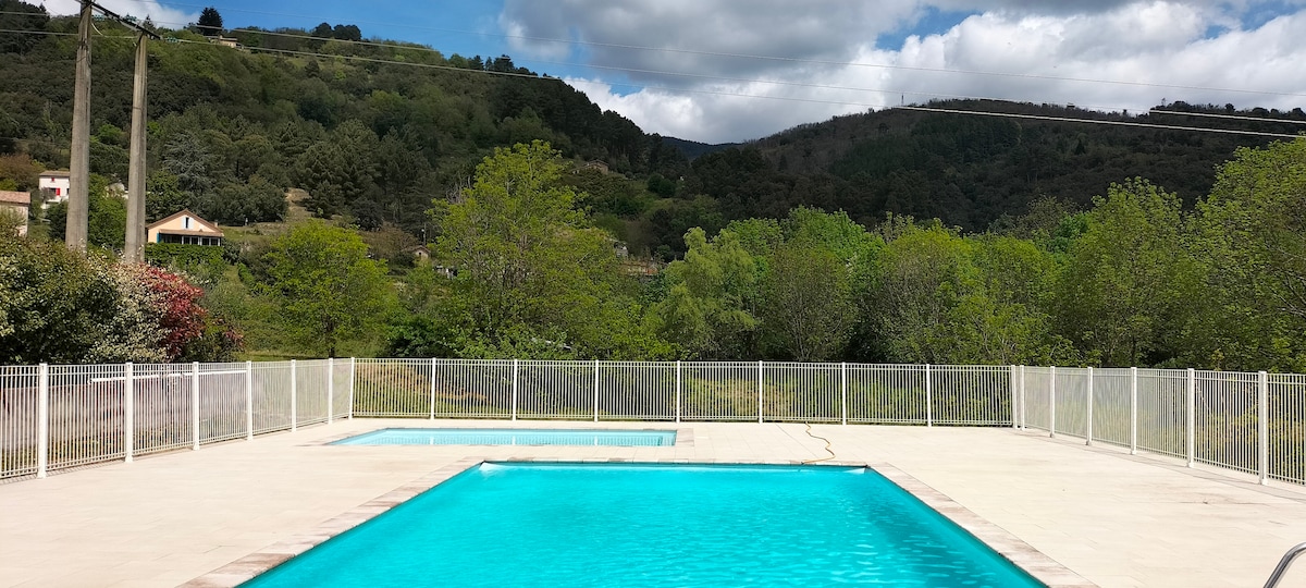 An outdoor swimming pool area is featured, with two pools surrounded by a white safety fence. Lush greenery and hills are visible in the background, under a partially cloudy sky. The serene atmosphere invites relaxation and leisure.