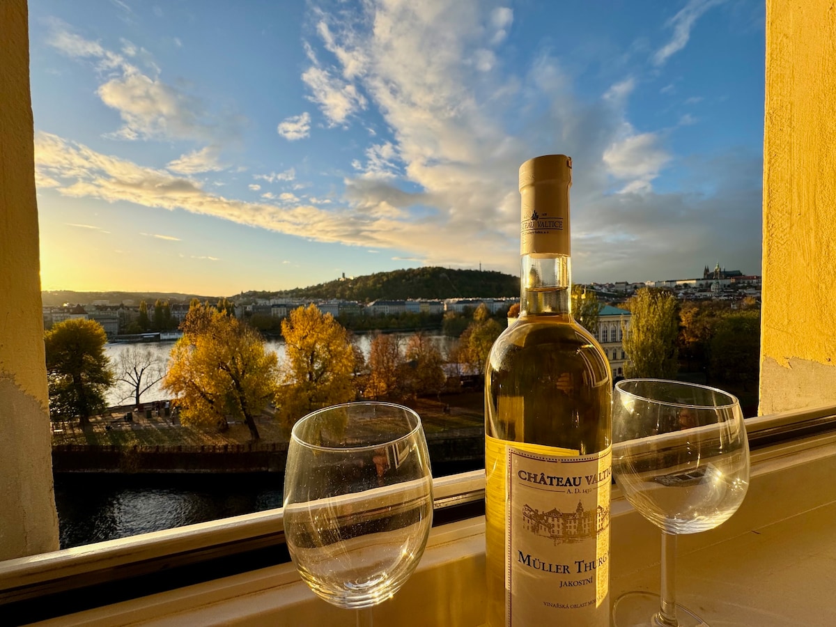 A bottle of white wine is positioned beside two elegant wine glasses on a window sill. Outside, a scenic view of the river and colorful autumn trees is visible, under a sky displaying soft blue and golden hues, suggesting a serene moment.