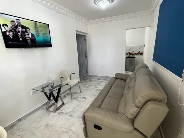 A living area is presented with a light-colored reclining sofa facing a mounted television. A glass table with a modern design accompanies a single chair. The walls are painted in neutral tones, and a doorway leads to another space, creating an open feel.