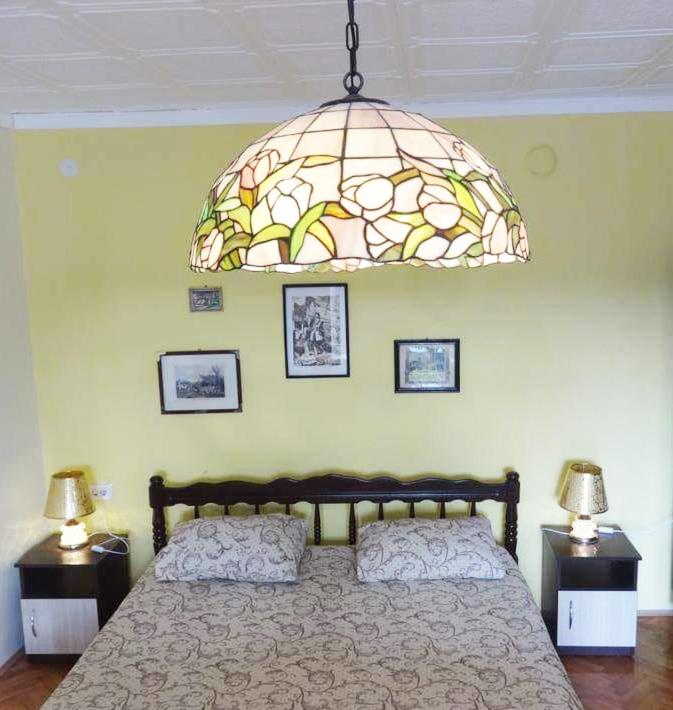 A bedroom features a spacious bed with a patterned coverlet. Two bedside tables flank the bed, each with a lamp. A large stained glass light fixture hangs overhead, and colorful framed photographs are mounted on the yellow walls.