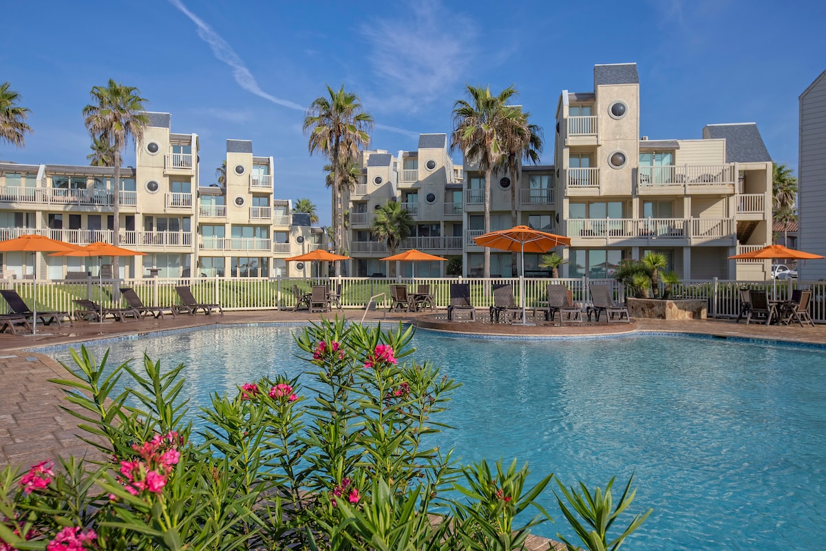 A clear view of a spacious pool area is presented, surrounded by lounge chairs and umbrellas. Lush greenery and vibrant flowers frame the pool, while palm trees provide a tropical ambiance. Multifamily condominiums are visible in the background, showcasing their modern design.