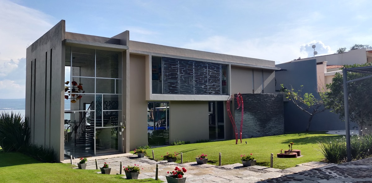 The exterior of a modern residence is depicted, showcasing a combination of stone and glass elements. Lush green grass surrounds the building, with neatly arranged flower beds bordering a stone pathway. Large windows provide a view of the lake, reflecting natural light.
