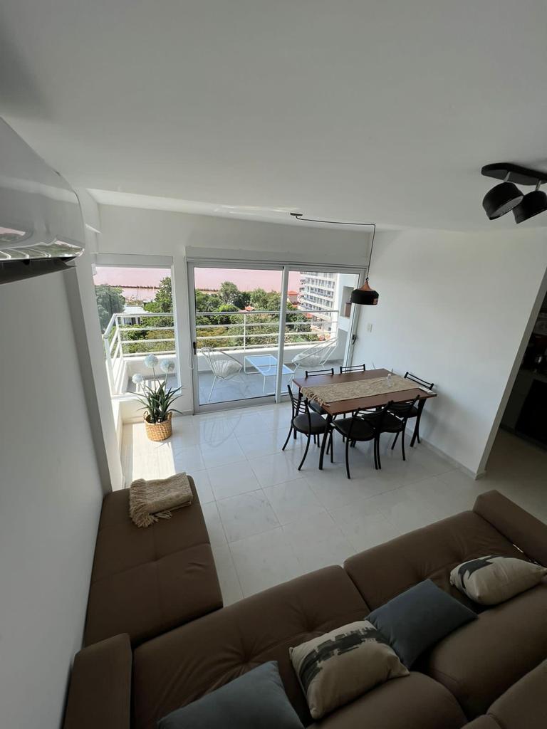 A light-filled living area showcases a spacious lounge with a comfortable sofa. A dining table with six chairs is positioned near large glass doors that provide beautiful views of the surrounding landscape and natural light.