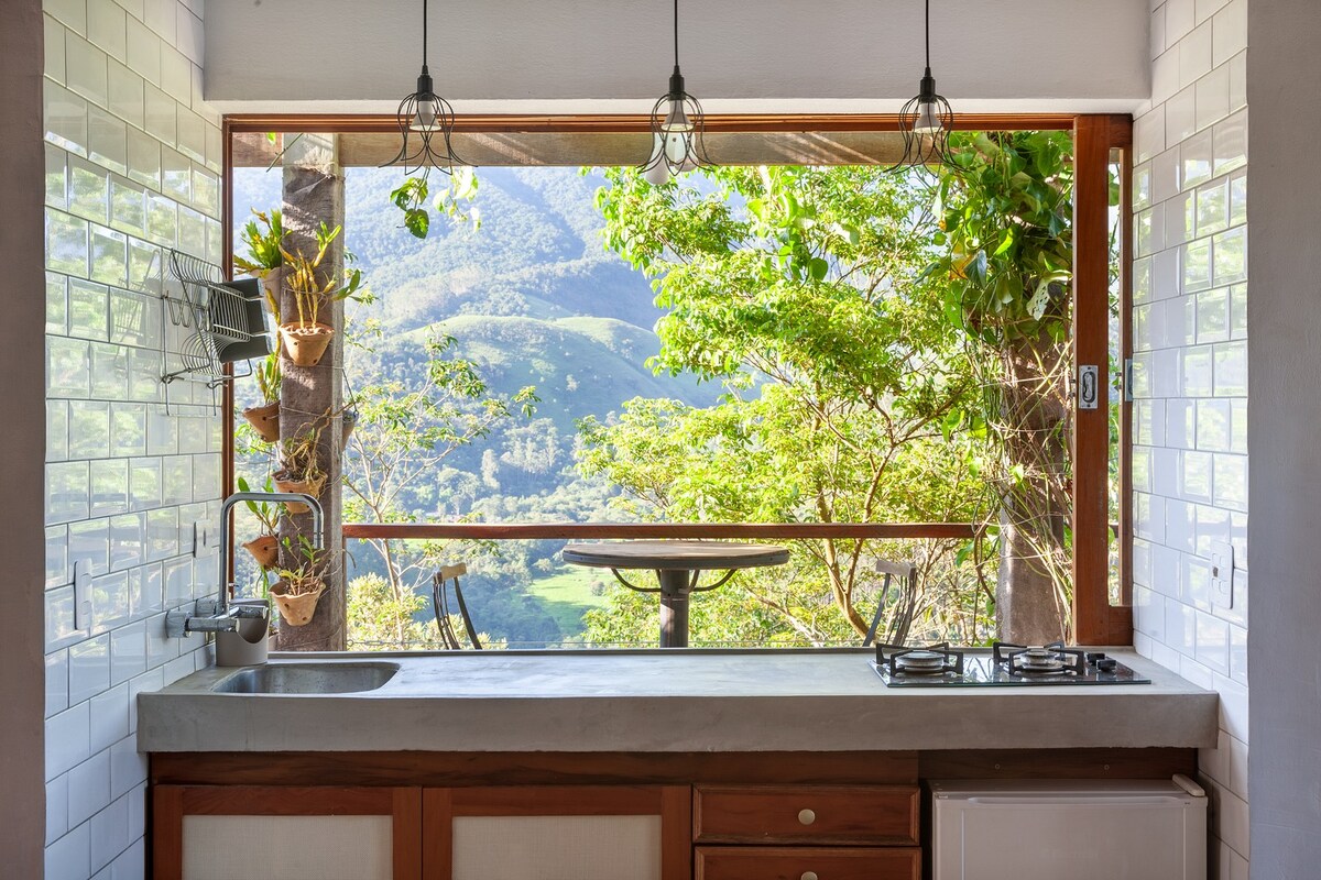 A kitchen area features a long counter with a sink and a stove, offering a view of the outdoors through large windows. Green foliage and mountains are visible in the background. Three pendant lights hang above the counter, enhancing the natural light in the space.