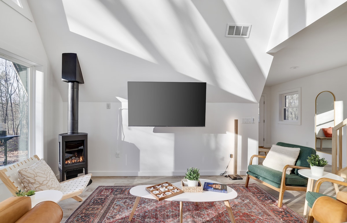 A bright living area features a cozy gas fireplace beside a large flat-screen TV mounted on a white wall. A modern coffee table sits on a patterned rug, complemented by stylish seating options, including a green chair and a minimalist white lounge chair. Natural light streams through expansive windows.