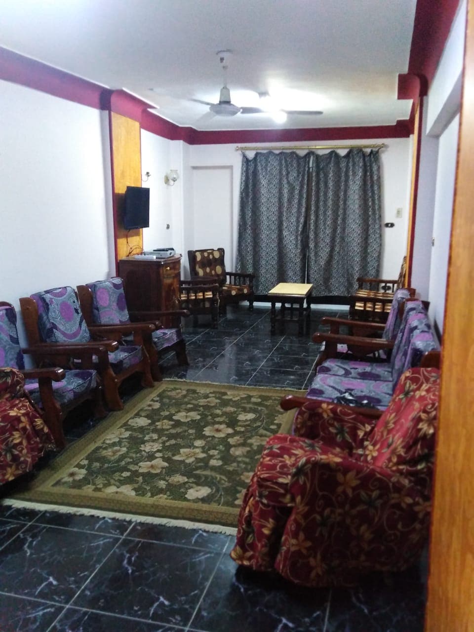 A spacious living area features comfortable seating arrangements with patterned chairs and a dark rug at the center. A wooden table is placed between seating, complemented by decorative curtains covering a window. A wall-mounted television adds to the entertainment options in the space.