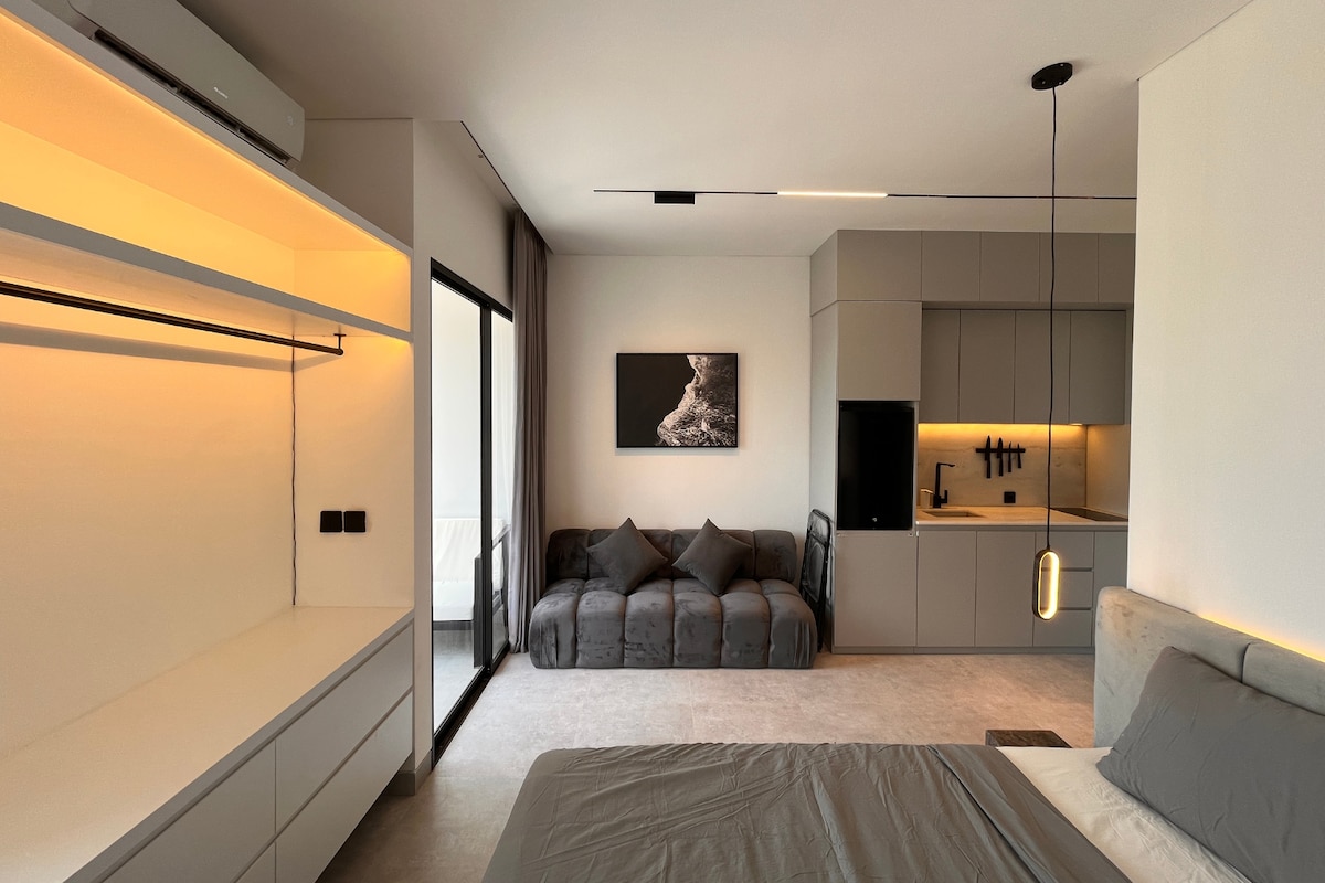A modern interior space features a spacious bed with neutral bedding and stylish recessed lighting. A comfortable couch sits against the wall, complemented by a sleek kitchenette with minimalist cabinetry. Large glass doors offer access to the balcony, enhancing natural light throughout the area.
