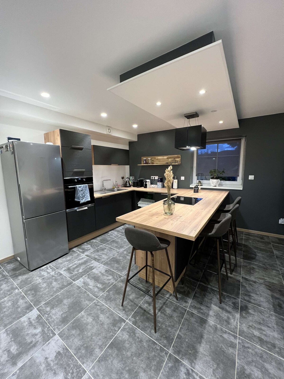 The modern kitchen features dark grey walls and stylish wood accents, highlighted by overhead lighting. A central island provides seating with three sleek stools, and appliances including a refrigerator and oven are visible. A window allows natural light to enhance the inviting space.