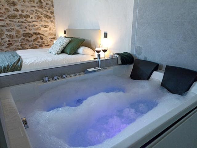 A relaxing jacuzzi tub is set against a stone wall, featuring bubbly water illuminated by soft blue lighting. In the background, a cozy bed is prepared with decorative pillows, and two glasses of wine are placed on a nearby nightstand.