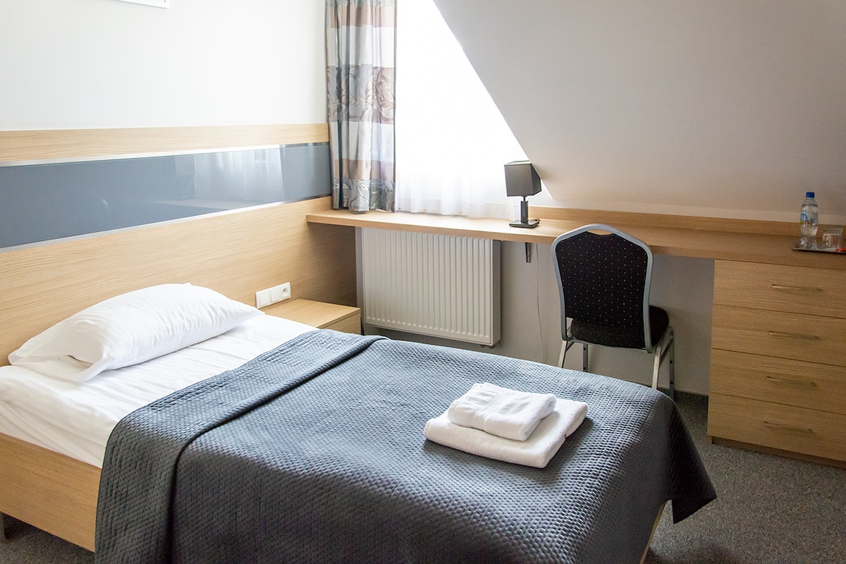 A well-lit guest room features a spacious bed with folded towels placed neatly on top. A desk with a chair is positioned against the wall, accompanied by a lamp. Natural light filters through the window, illuminating the simple, modern decor.