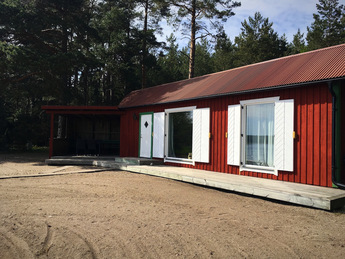 A red cottage with a sloped roof is situated among tall trees. Large glass doors allow natural light to enter, while framed shutters add a touch of charm. A wooden deck provides a seamless transition between the interior and sandy outdoor space.
