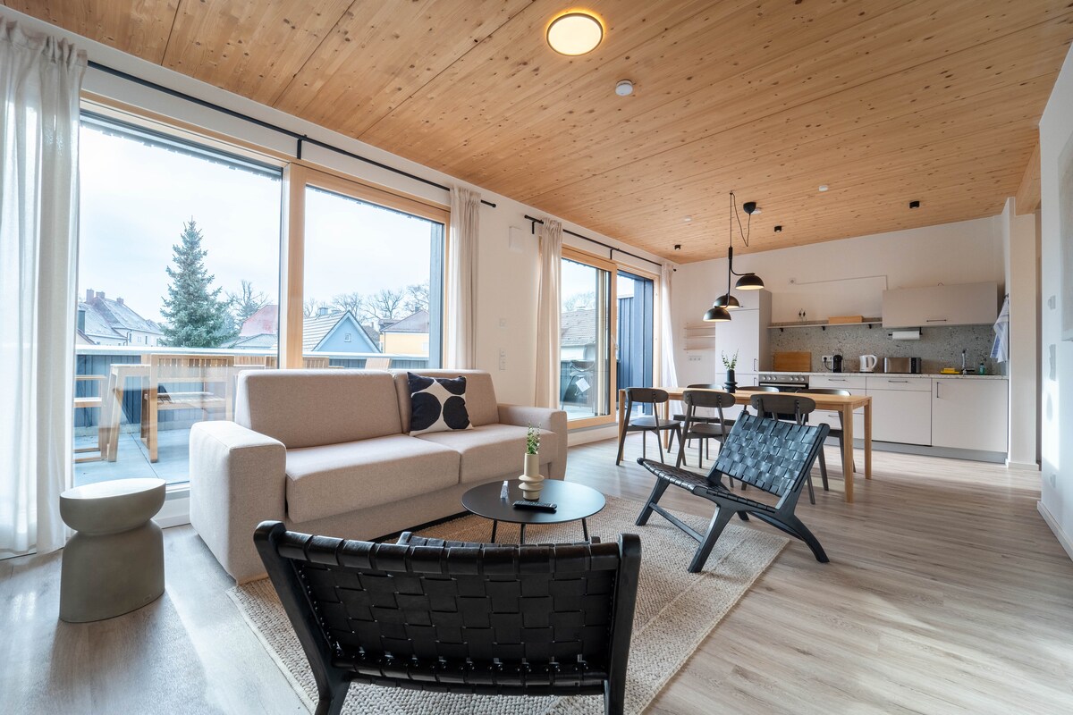 A spacious living area features a light-colored sofa, accompanied by two black armchairs and a low round table. Large windows illuminate the space and provide access to the terrace. A modern kitchen is visible in the background, designed with clean lines and functional appliances.