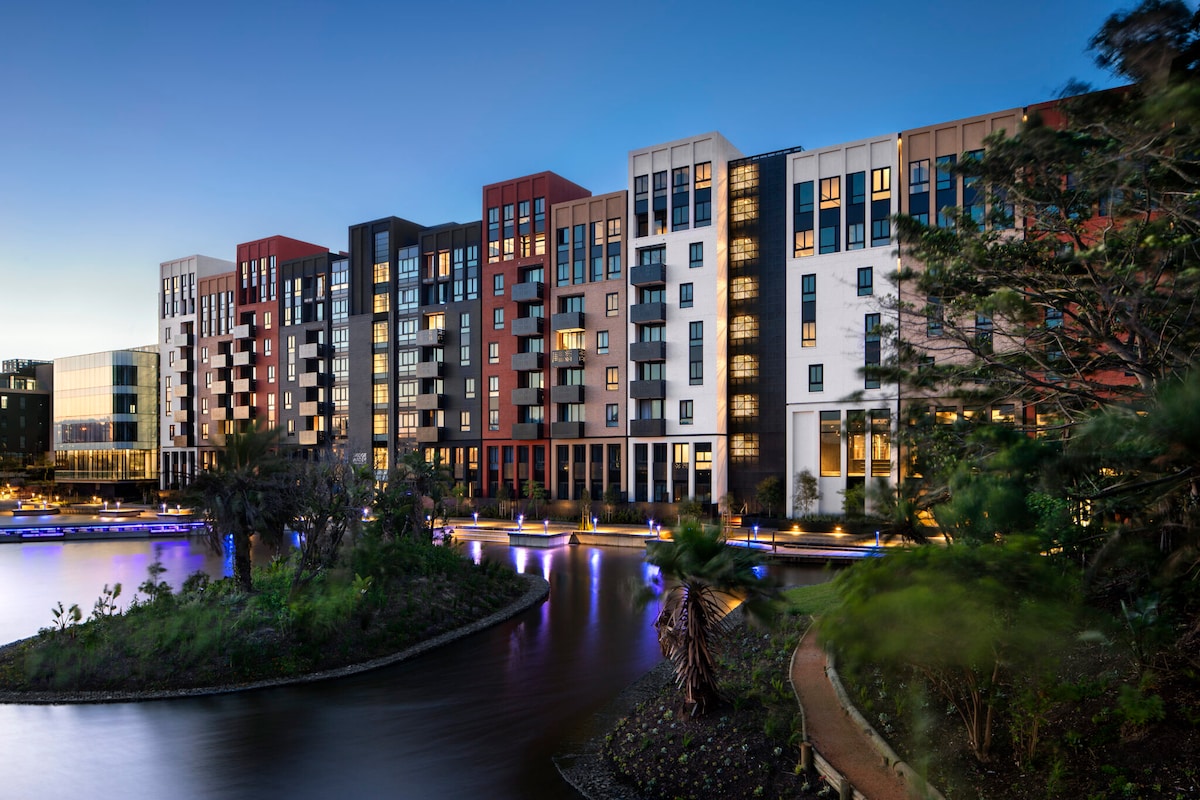 The multi-story apartment complex features a modern architectural design with a mix of colors, including red, black, and white. Surrounding greenery and a water feature add to the aesthetic, while soft lighting enhances the evening ambiance. Balconies provide outdoor space from many units.