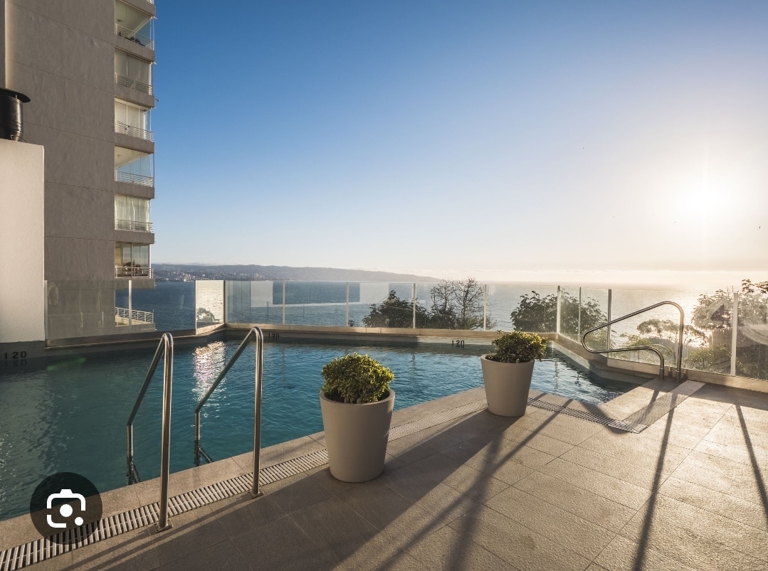 A serene swimming pool is framed by minimalist white walls, offering a panoramic view of the sea. The area is bathed in warm sunlight, with potted plants placed by the poolside. Glass panels provide a clear barrier, enhancing the overall calmness and elegance of the space.
