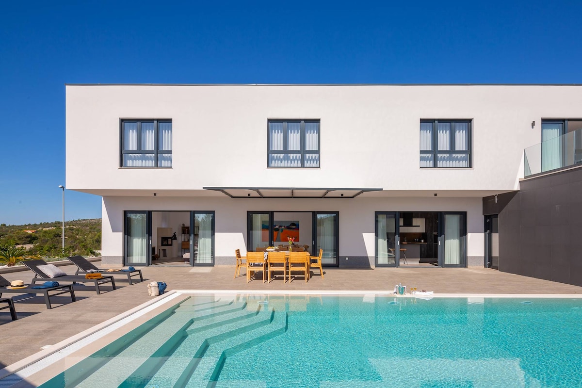 The exterior of the villa showcases a modern design with large floor-to-ceiling windows and an expansive patio area. A private pool reflects the clear blue sky, surrounded by sun loungers and an outdoor dining table. The space invites outdoor living and relaxation.