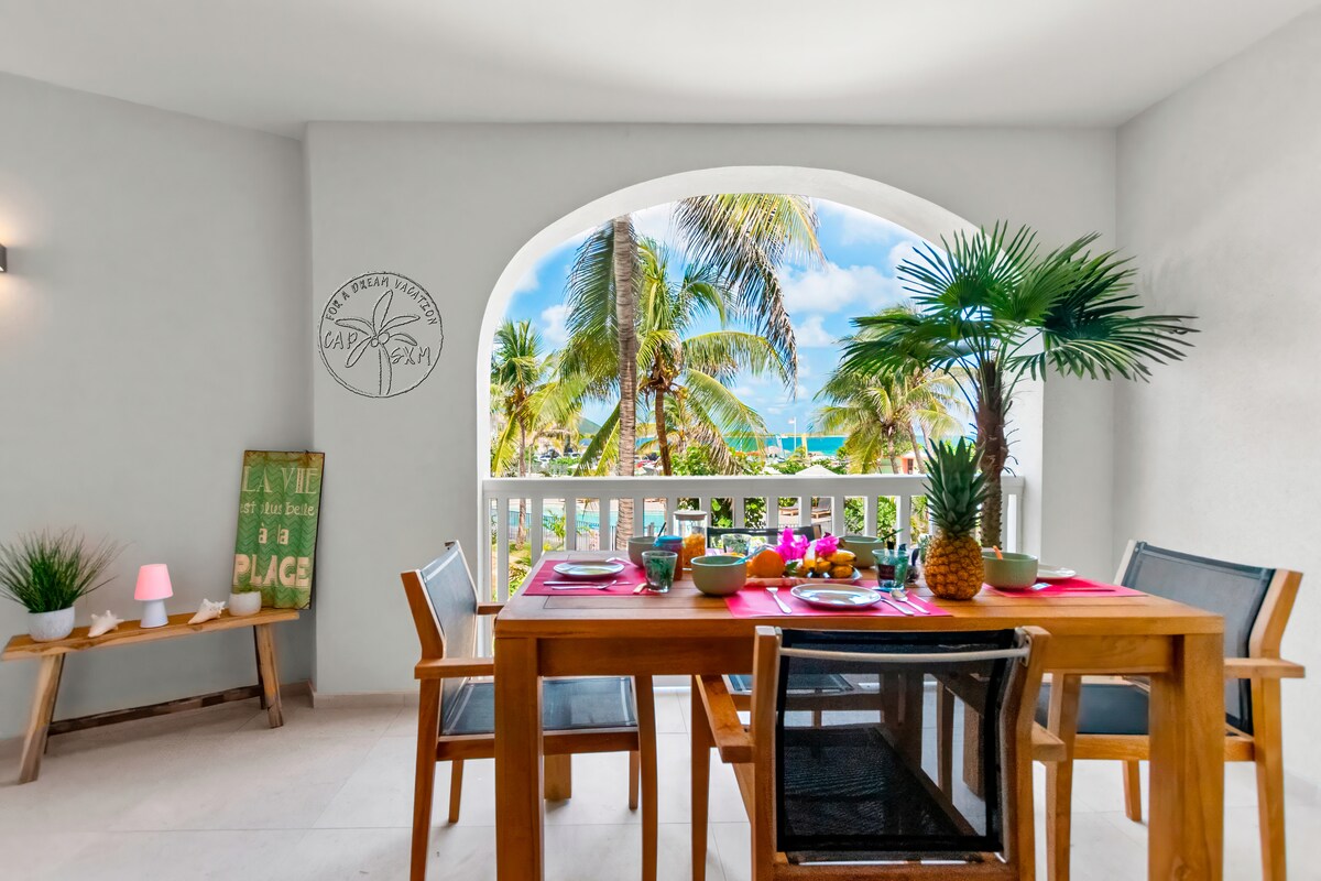 A dining area features a wooden table set with colorful dishes. Large windows allow natural light and provide views of palm trees and the ocean. Decorative plants add a touch of greenery, creating an inviting space for meals and relaxation.
