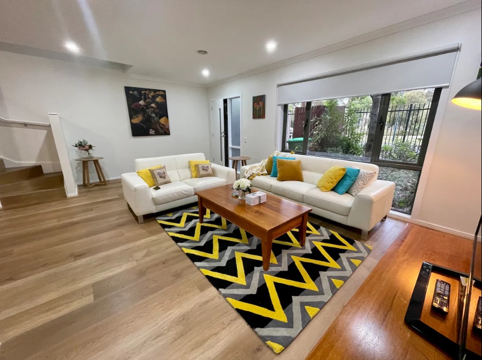A modern living area is presented with two white sofas adorned with yellow and blue cushions. A wooden coffee table is centered on a patterned rug that features bold black and yellow lines. Large windows invite natural light, revealing views of the greenery outside.