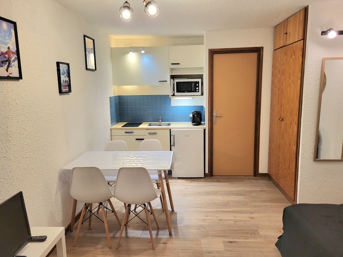 A modern kitchen area is visible, featuring a microwave and a compact stovetop. A dining table with four chairs is positioned nearby. Light-colored wooden floors enhance the bright atmosphere. The walls display framed artwork while a door leads to the hallway.
