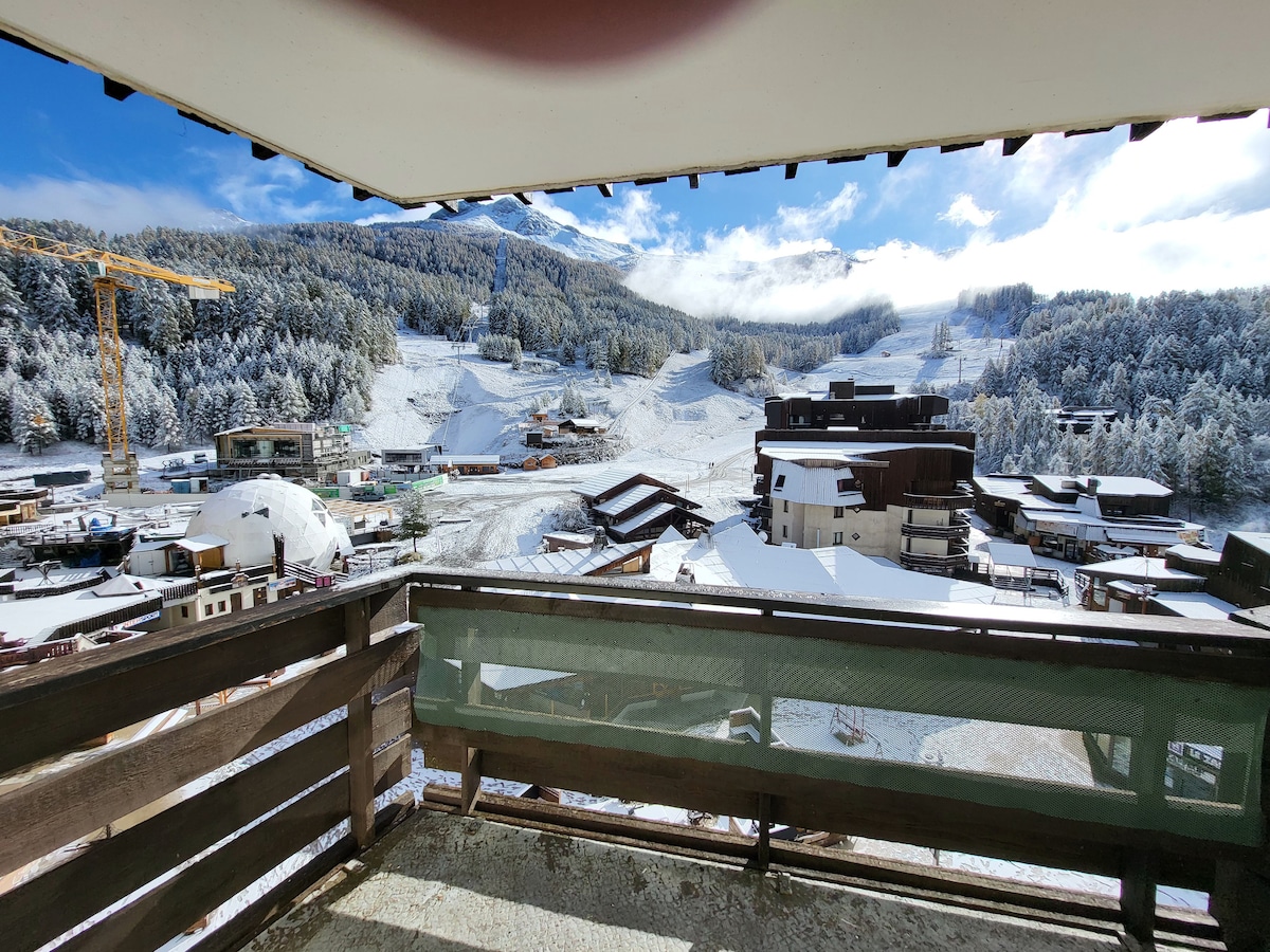 A balcony offers a wide view of a snow-covered landscape, including ski slopes and buildings. Lush trees are visible in the background against a clear blue sky with scattered clouds, creating a serene winter scene.