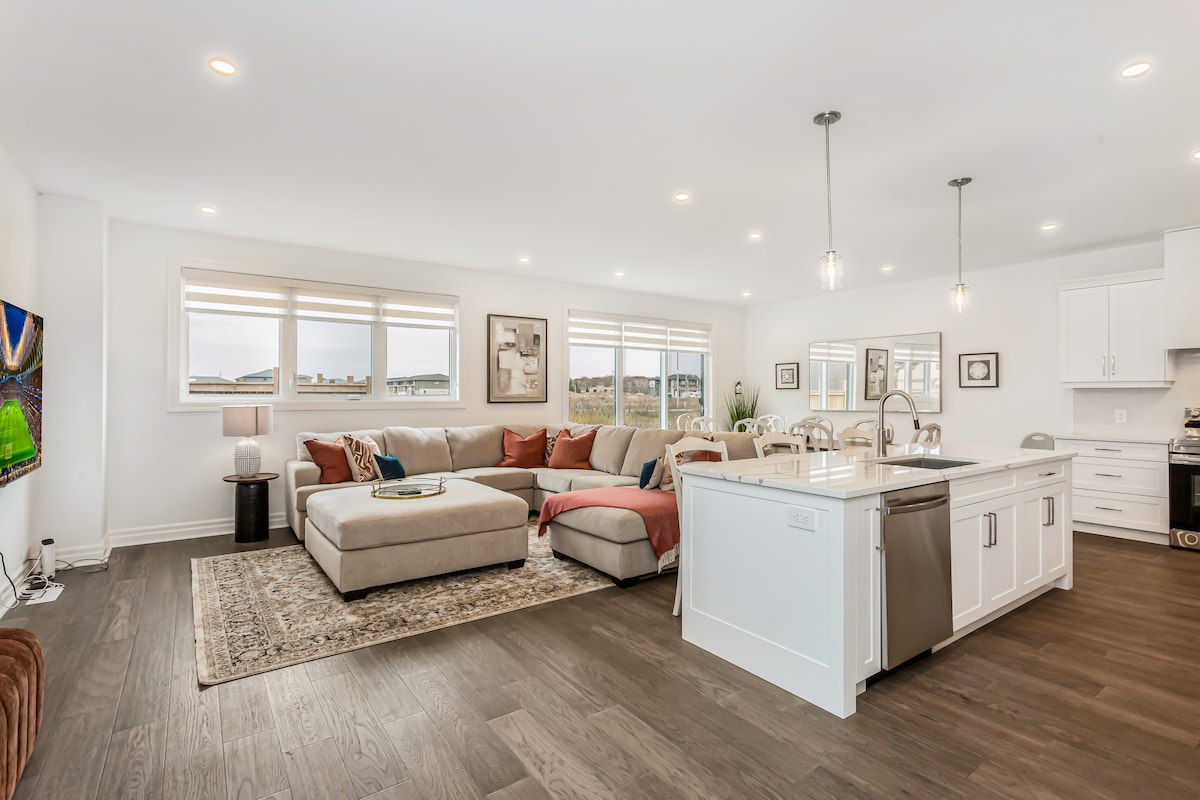 An open-concept living area features a spacious sectional sofa adorned with decorative cushions, complemented by a light rug. The kitchen island includes modern bar stools, and large windows allow natural light to brighten the space, providing a welcoming ambiance for relaxation and dining.